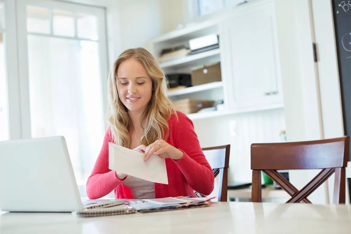 A women wearing a red sweater holding an envelope in her hand and has a laptop before her.