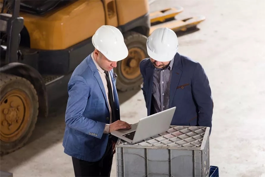 Two men wearing a suit and a construction safety helmet and has a laptop before them and discussing.