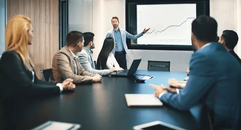 A man giving a presentation in a meeting room with a projector showing a graph. There are at least 7 people attending this meeting.