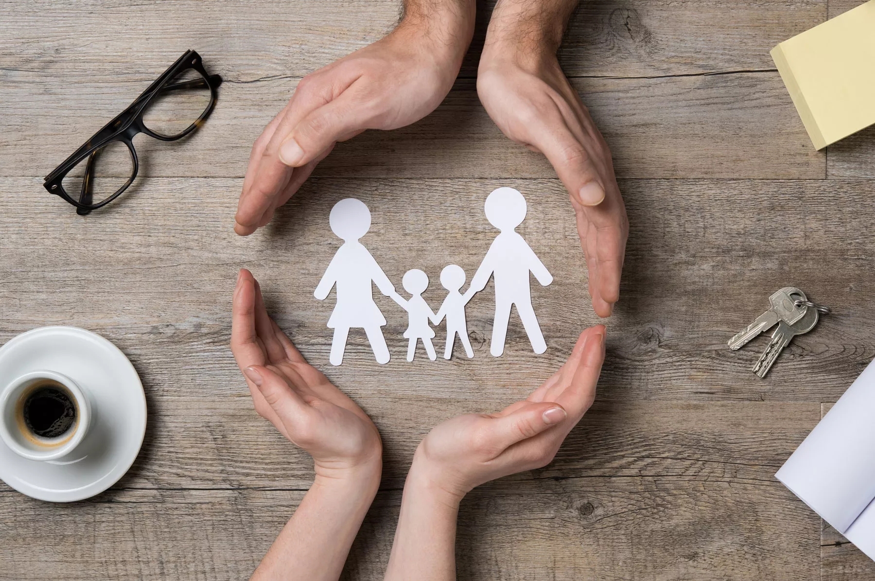 picture showing hands wrapped around picture of a family of four and has keys, coffee cup and glasses on the table.