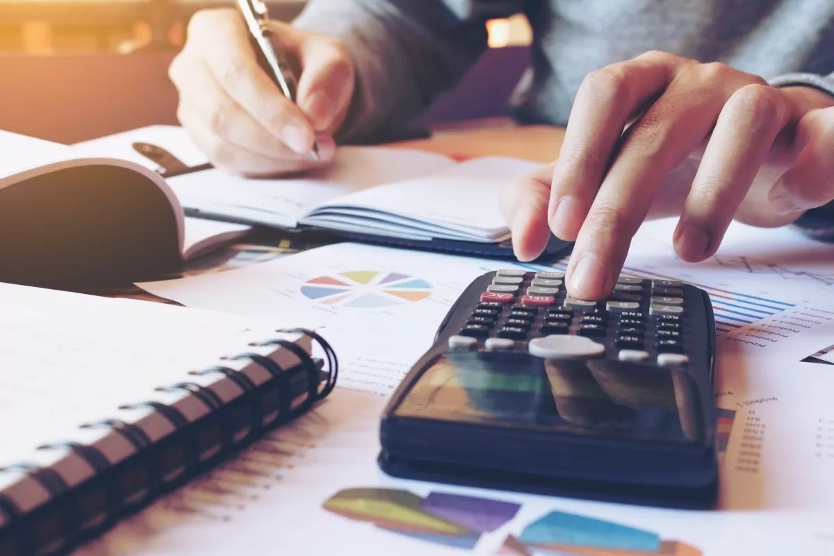 A person's hand using a calculator with many graphs and data opened on the table in front of him.