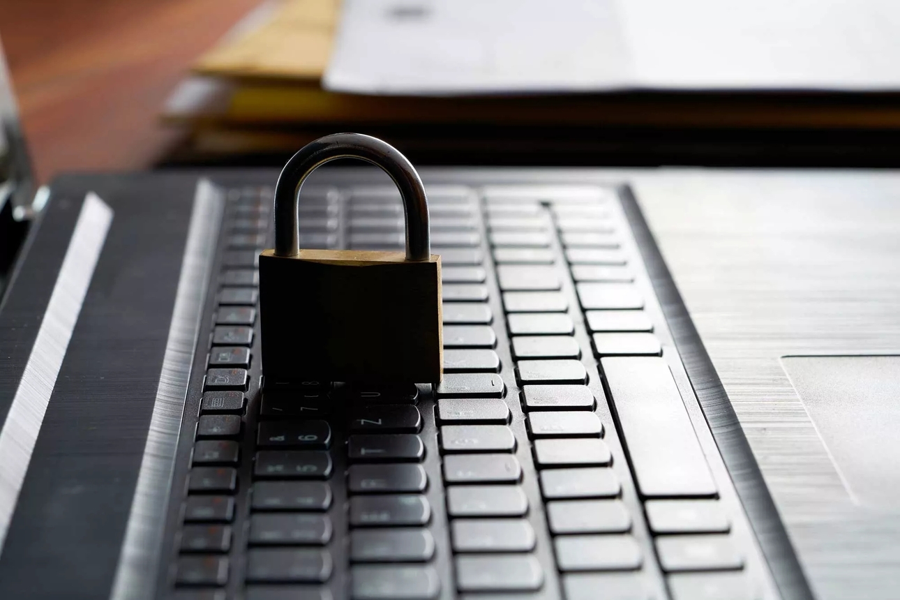 A sturdy padlock sitting on an open laptop's keyboard. The scene is viewed from the laptop's side and illuminated from behind.