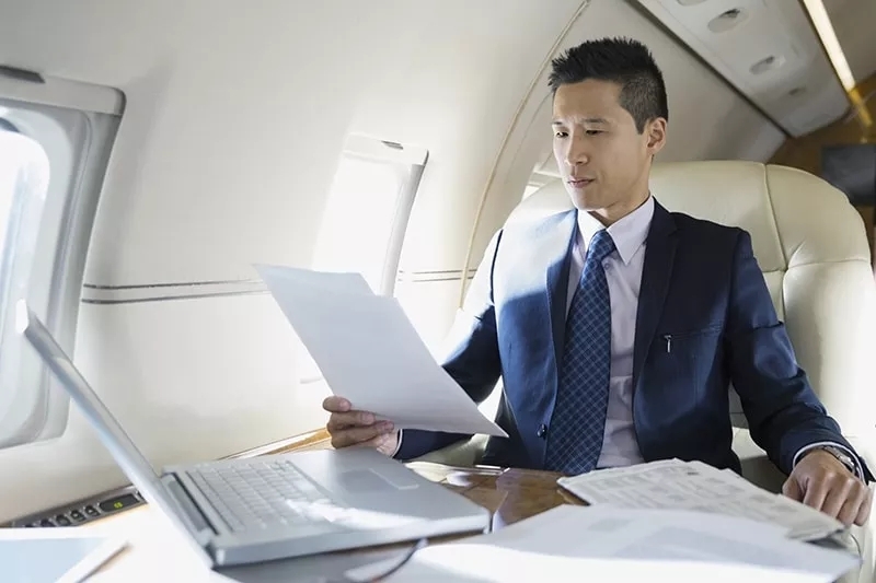 A man on an aircraft in flight, with papers and a laptop on the table in front of him.