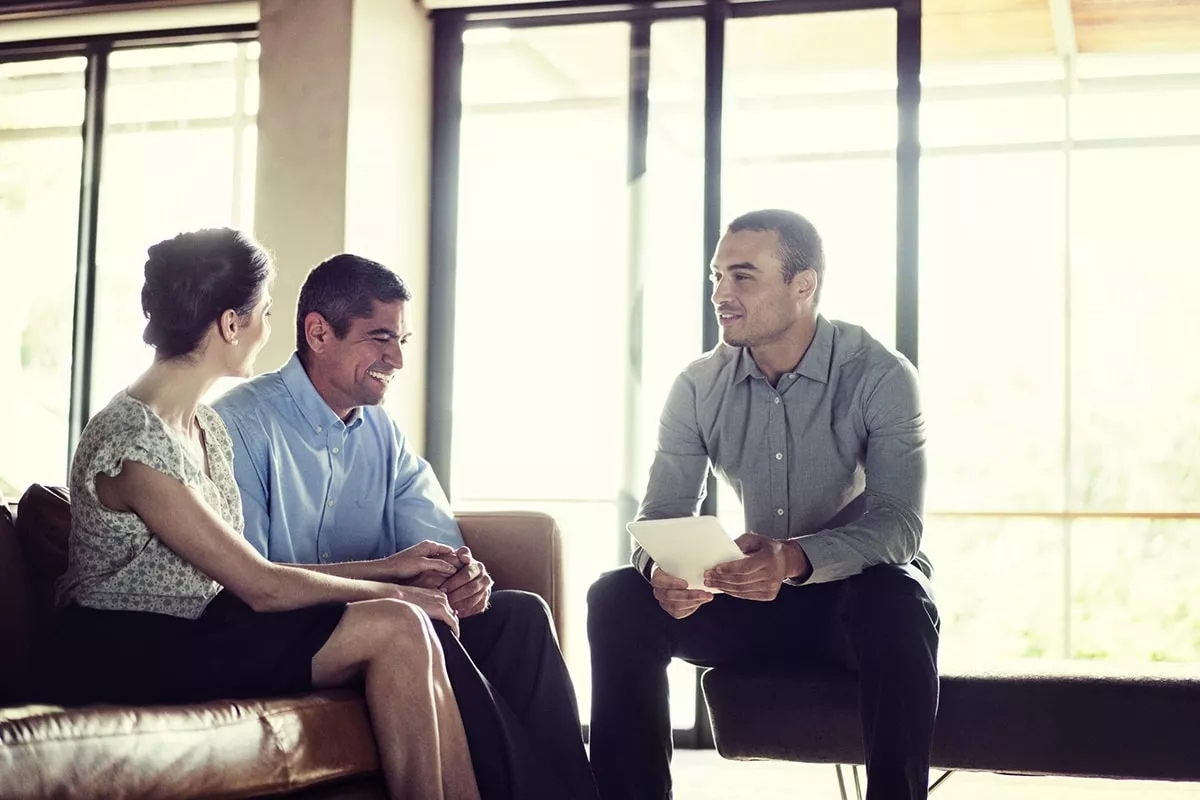 A couple in a meeting with another man with papers in his hand. They are all smiling in conversation.