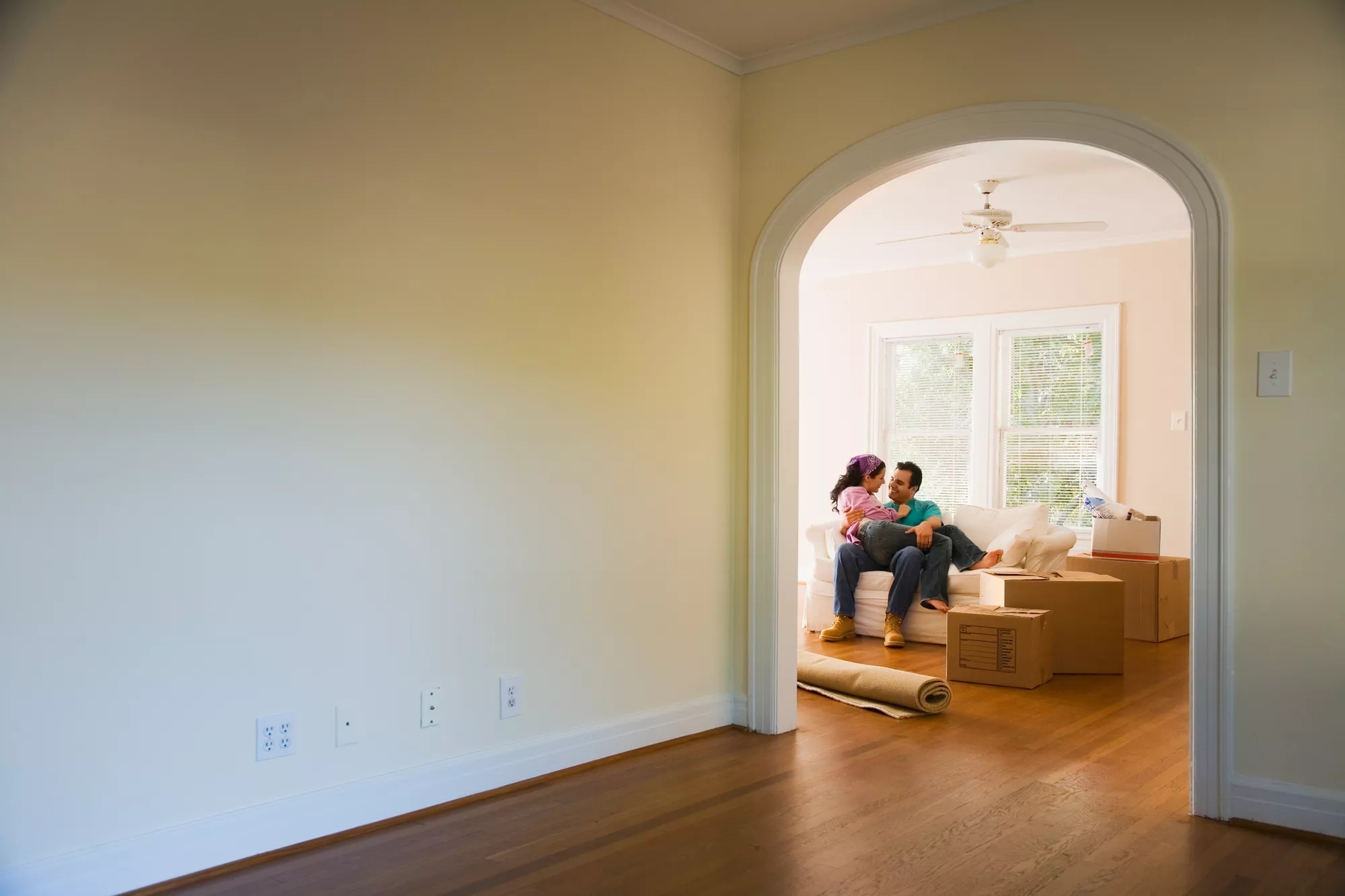 Two people sitting together on a couch through a doorway in a mostly empty hardwood house. The scene is bright and uplifting.