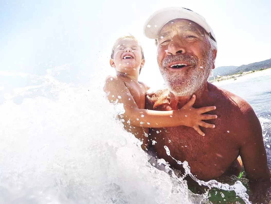 A grandfather holding his son amidst a cresting wave onto them, playfully splashing them in the chest.