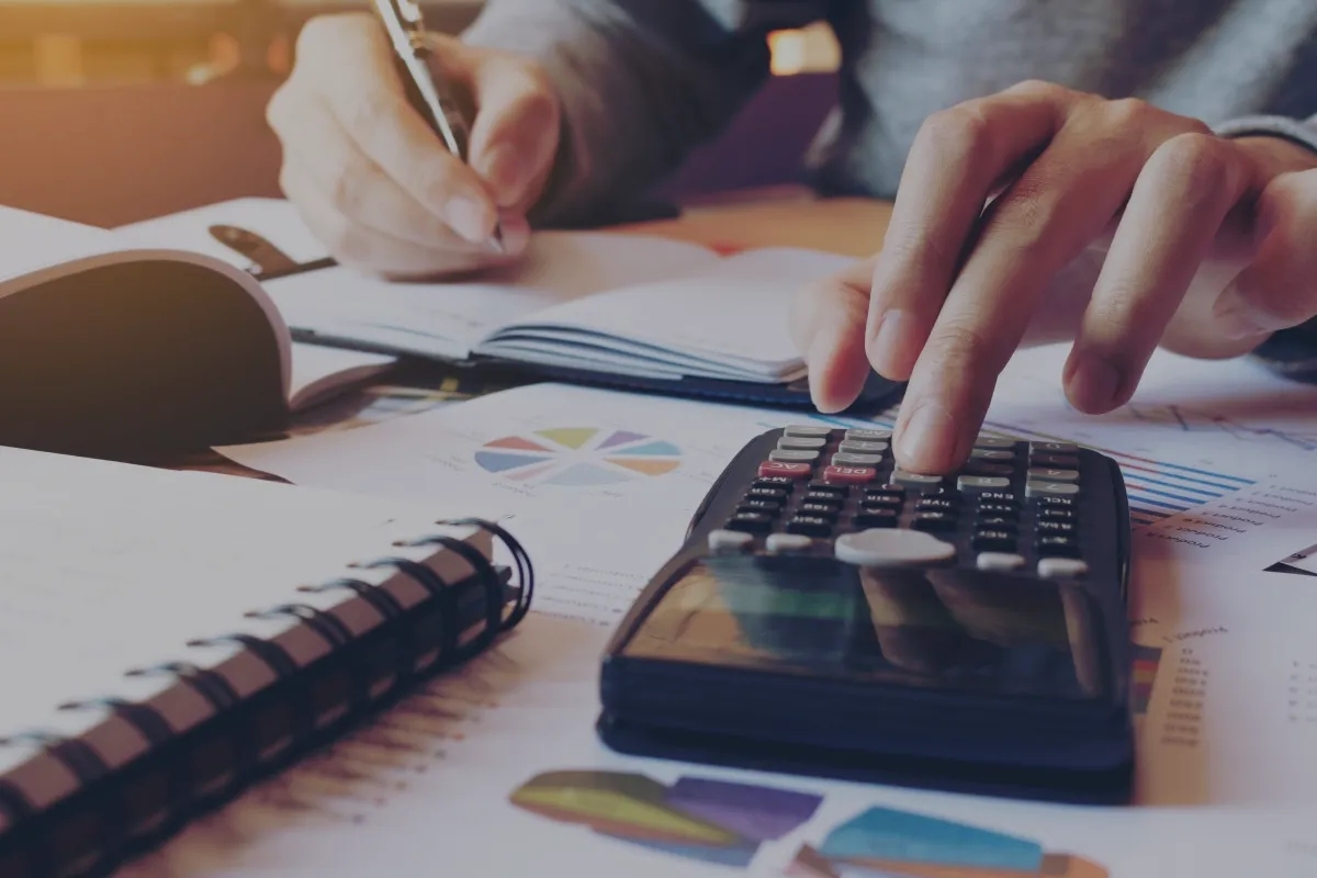 A person's hand using a calculator with many graphs and data opened on the table in front of him. A person's hand using a calculator with many graphs and data opened on the table in front of him.