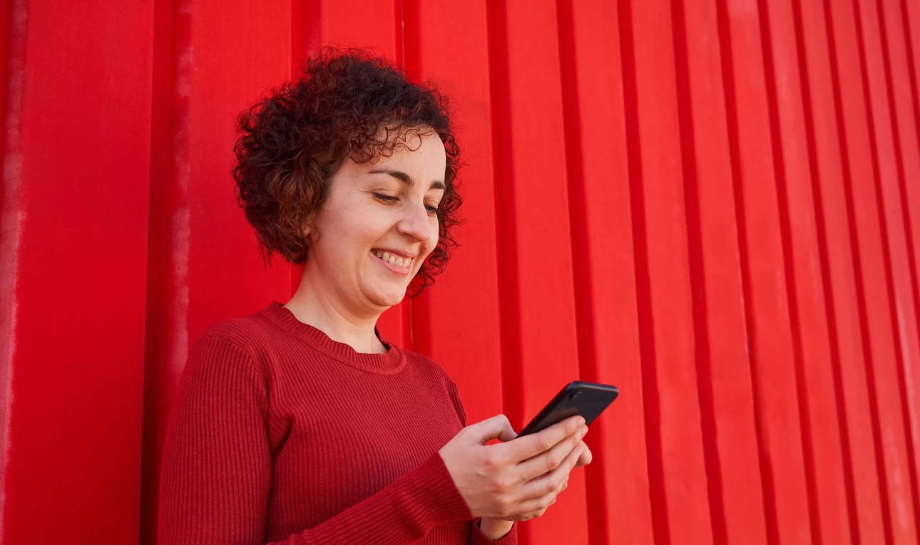 A smiling woman looking down at her phone, leaning against a red, slatted wall.