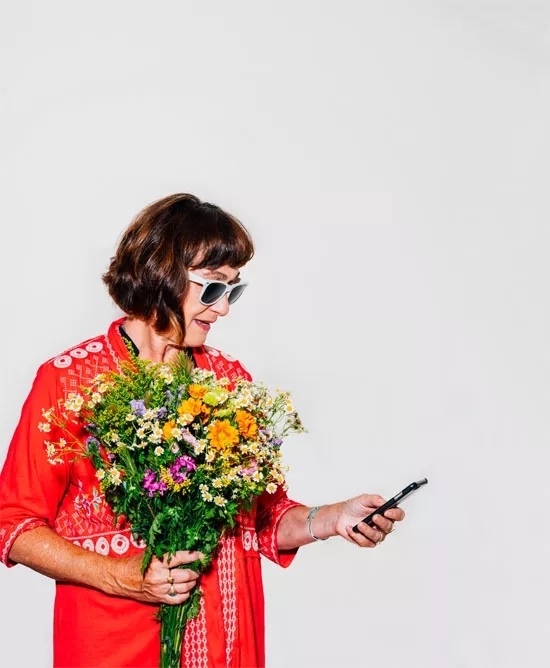 A woman in a vibrant red shirt holding a bouquet of flowers in one hand and a smartphone in the other. She's wearing sunglasses.