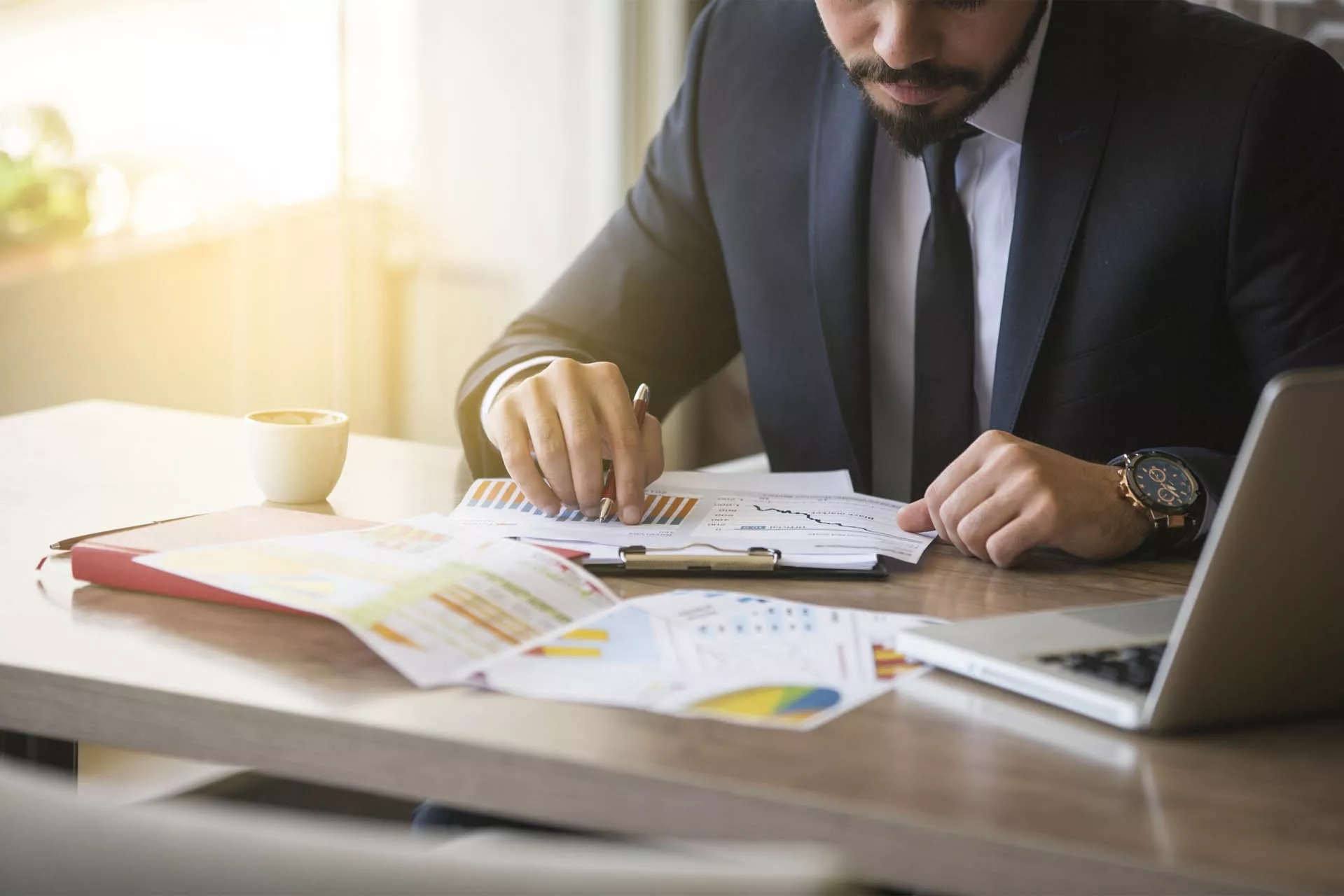 A person sitting at a desk with an open laptop and many analytical papers with charts and information on them on the table in front of him.