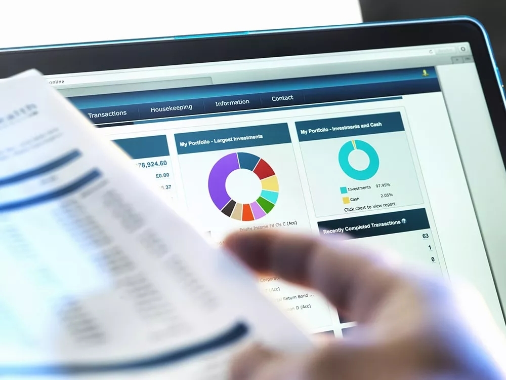 A dashboard on a computer, over the shoulder of a person near to the camera holding a paper spreadsheet.