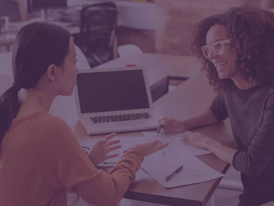 Two women discussing an open laptop on the table between them.