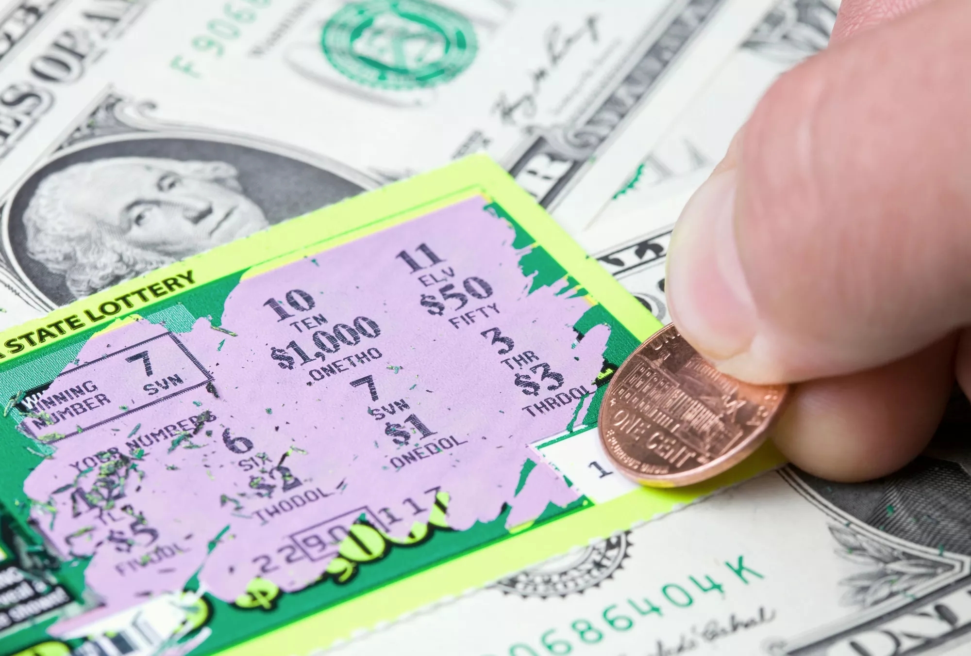 A lottery ticket being scratched with a penny, on top of spread out one dollar bills on the table below.