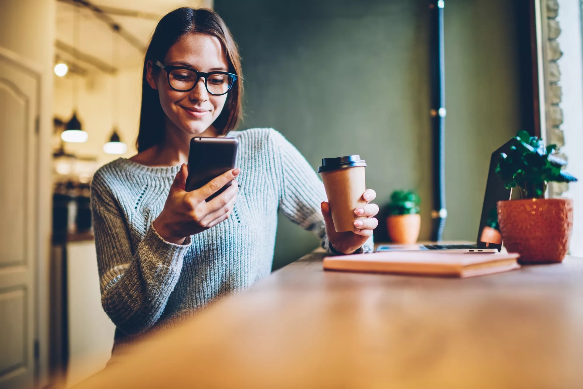 A woman leaning back in her chair at a coffee shop. She is smiling at her phone, with a coffee in the other hand. A woman leaning back in her chair at a coffee shop. She is smiling at her phone, with a coffee in the other hand.