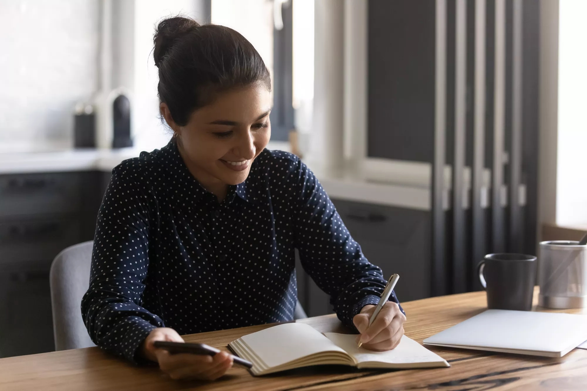 A smiling person writing in an open book, with her phone in the other hand.