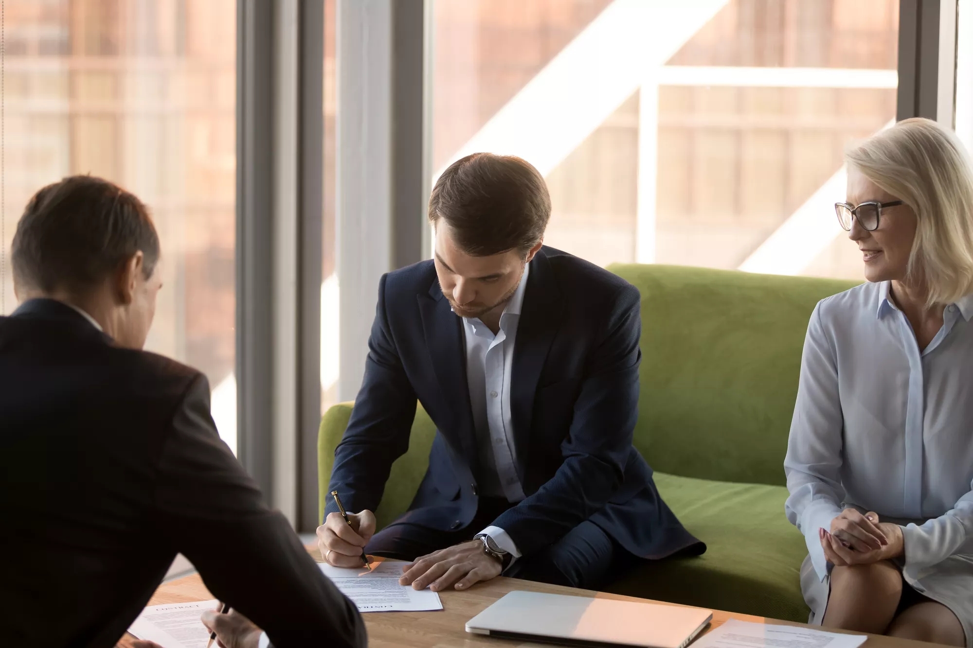 Business man signing paperwork in a meeting of three individuals.