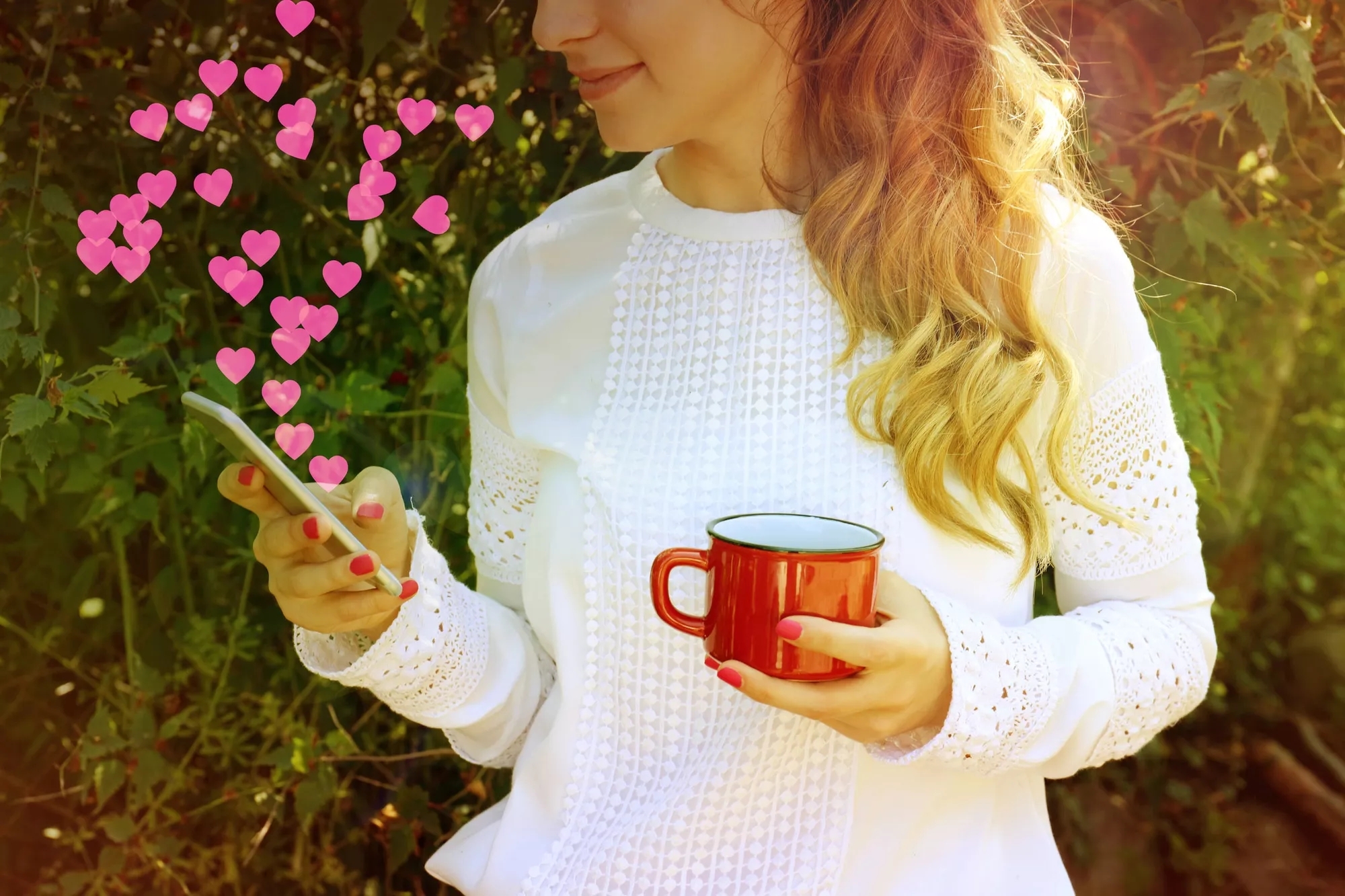 A woman is outside in front of some bushes, messaging on her phone, with heart emojis flying out, holding a mug in the other hand.