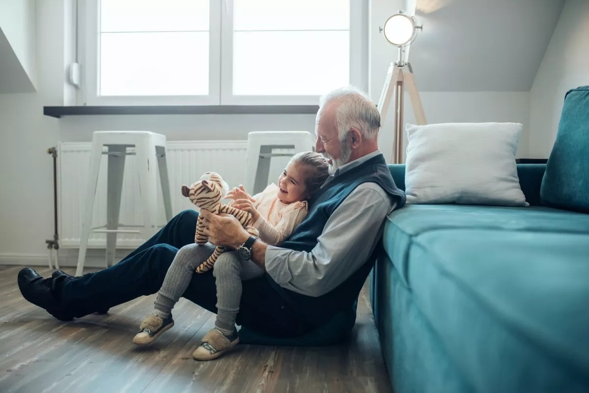 A grandfather shows his granddaughter a stuffed tiger on the floor against the couch.