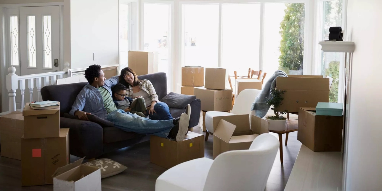 A family moving in, relaxing on couch. Unopened cardboard boxes are scattered around.