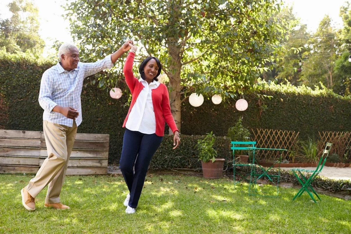 A husband twirls his wife outdoors to an unknown song. The day is bright.