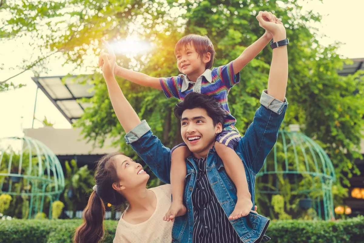 A child sitting on their father's shoulders with the mother looking from behind him smiling. The trees behind them are backlit with a soft sunset.