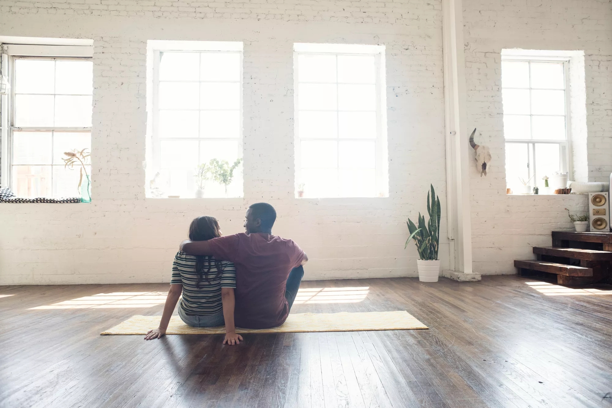 Two people sitting on a yoga mat in an emtpy room on the hardwood floor. They're looking jubilantly out the window towards the bright sky across from camera.